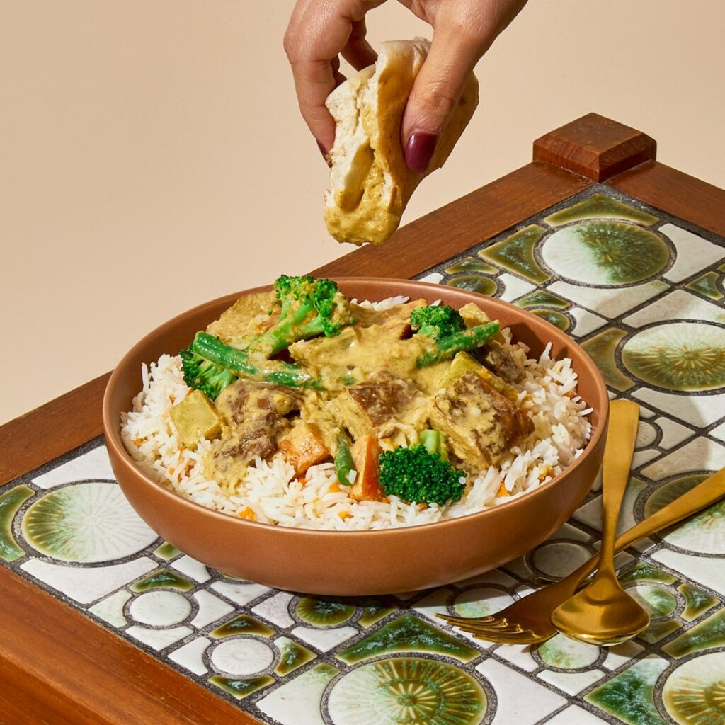 A bowl of Thai-style yellow curry on a bed of rice sits on a small table. A hand is just in shot holding some roti bread above to dip in. 