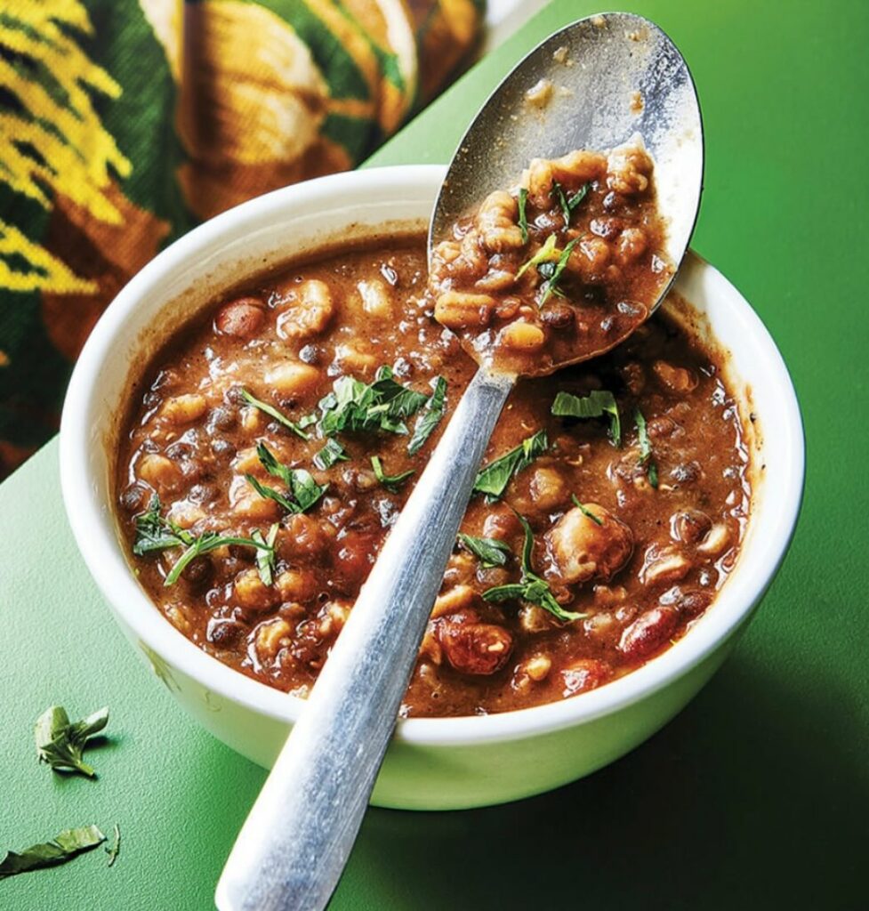 A bowl of bean soup sits on a green bench. A spoon with some soup on it balances on the top of the bowl.