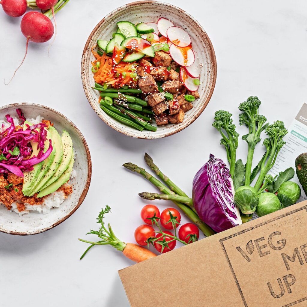 An overhead shot of two different vegan dishes next to a meal kit delivery box, with its ingredients splayed out.