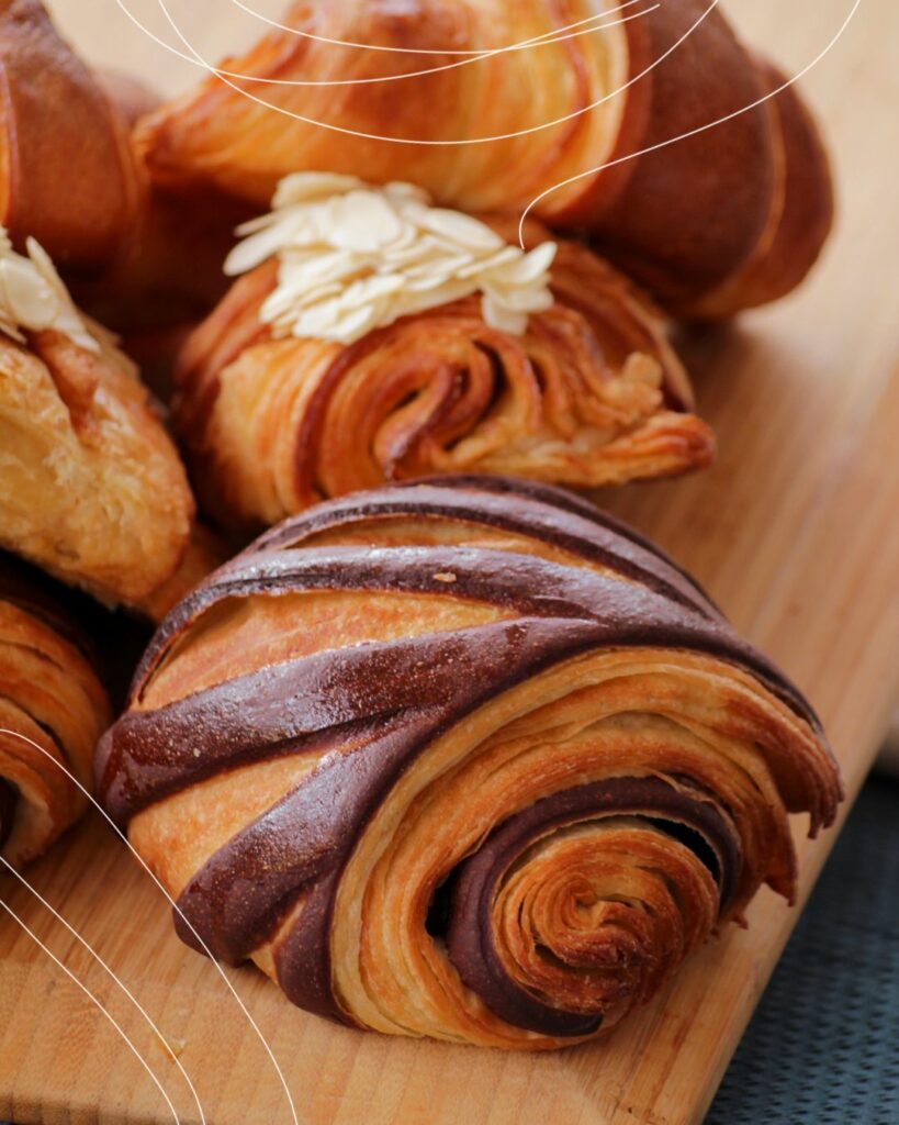 A close up image of a pain au chocolate with almond croissants piled up behind it.