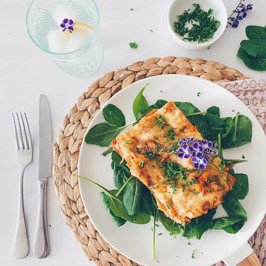 An overhead image of a vegan lasagne on a bed of spinach at a table setting. It is decorated with edible purple flowers.