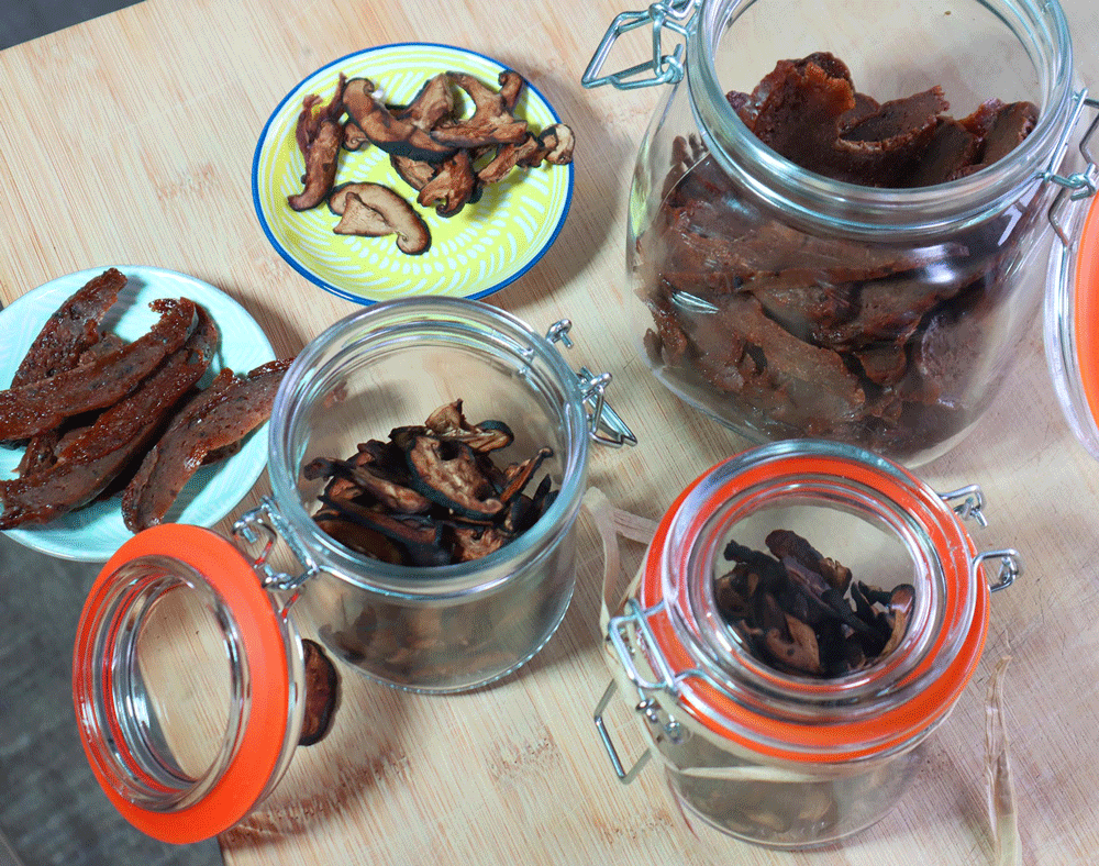 Several glass jars and plates sit on a bench full of a beef-style mushroom jerky.