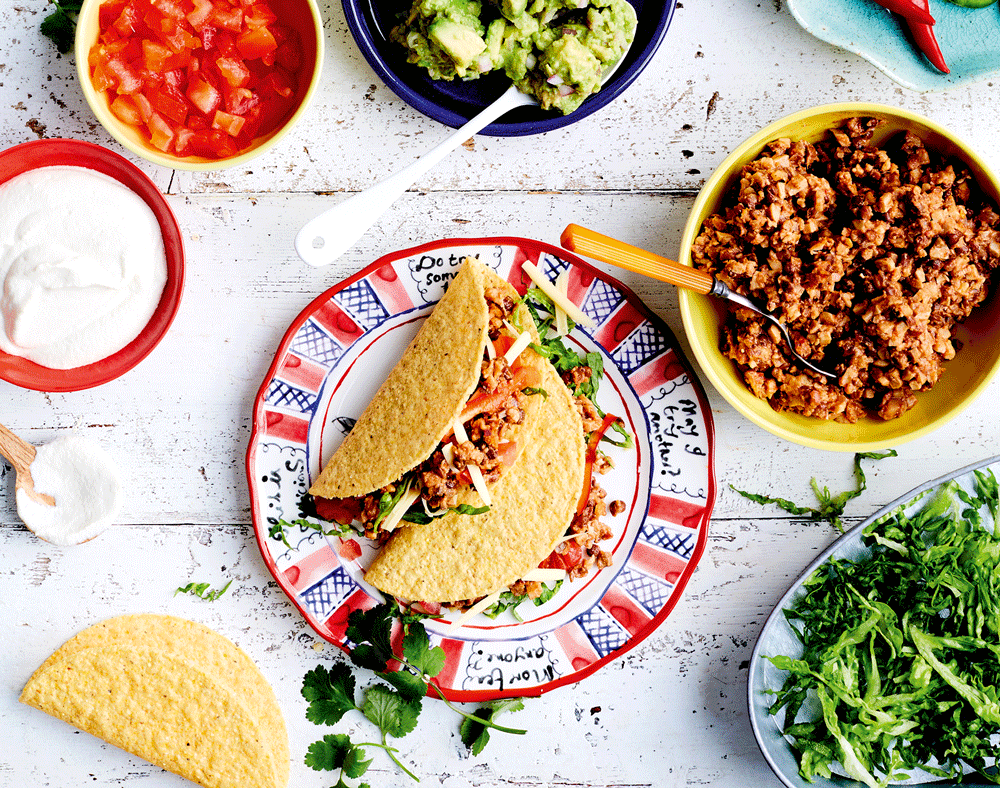 An overhead image of crispy taco shells filled with a lentil and walnut beef-style mince. The plate sits on a white bench surrounded by colourful additions including guacamole, diced tomato, sour cream, chilli and greens.