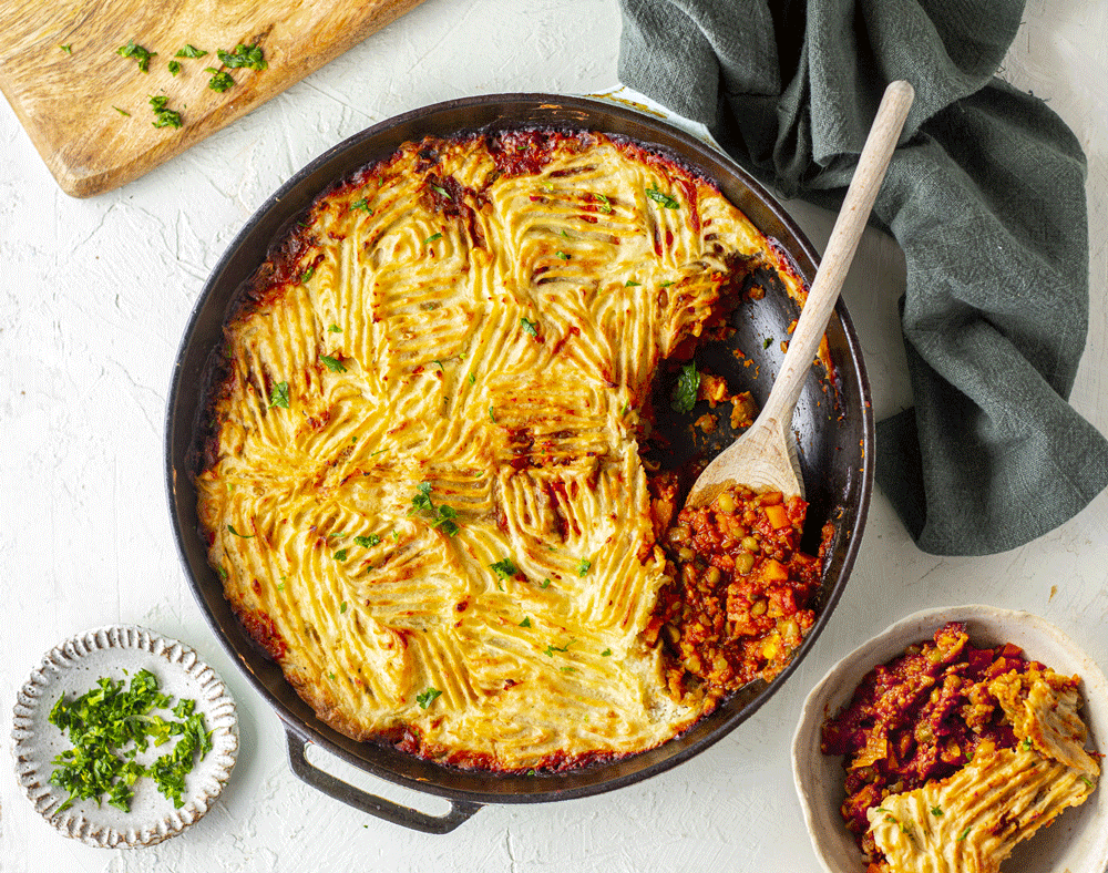 An overhead image of a skillet filled with Shepherd's pie. A slice is taken out and sits in a bowl beside, showing the delicious filling of hearty veg and lentils.