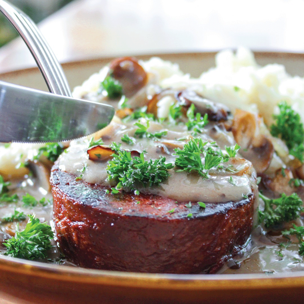 A round of beef-style steak is about to be cut into by a knife. It's topped with a creamy jus and garnished with greens. A pile of mash sits in the background.