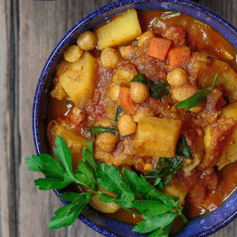 An overhead close-up image of a blue bowl filled with Moroccan Tagine. It's laden with vegetables and garnished with a side of parsley.