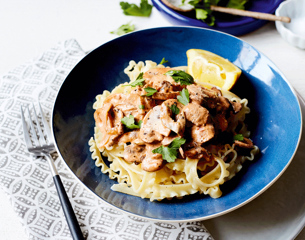 A blue bowl full of pasta with a rich mushroom stroganoff, served with a wedge of lemon.