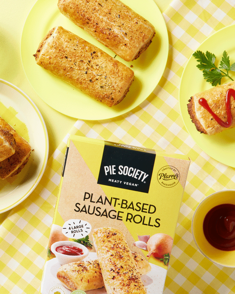 A bright yellow and white chequered tablecloth sits underneath several plates displaying Pie Society's plant-based sausage rolls. Some packaging sits in shot. They look like absolute perfection!