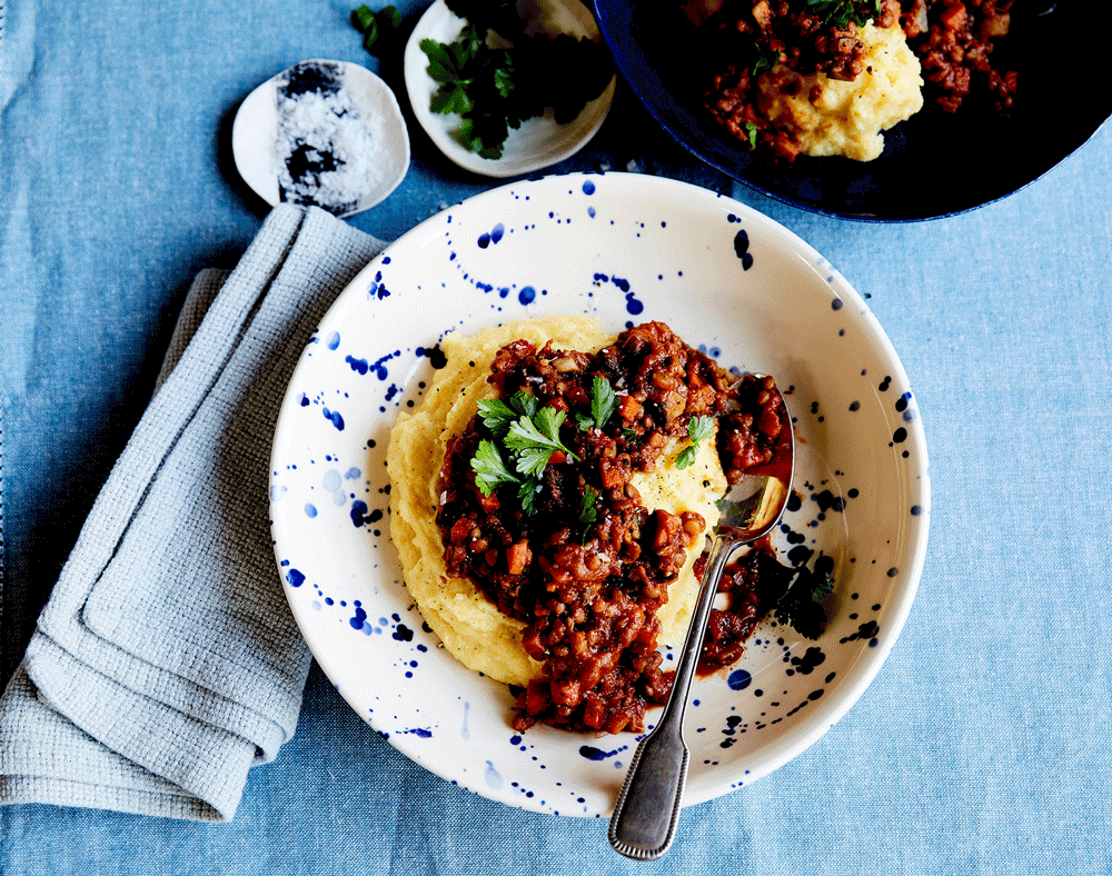 A rich lentil bolognese sits atop a bed of creamy polenta. A blue napkin sits to the side, slighty lighter in colour than the blue of the tablecloth.,