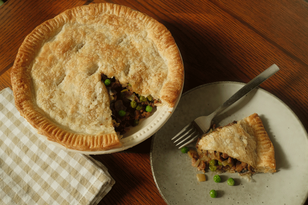 A full pie with flaky pastry sits on a bench next to a chequered tea towel. A slice of the pie sits on a plate to the side, with a beefy looking filling with peas.