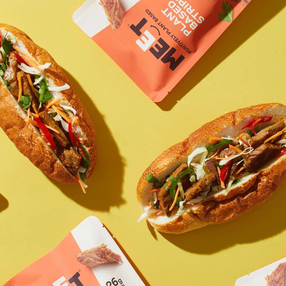 Two Bahn Mi style rolls sit on a bright yellow surface, made using Meet Co's plant-based strips. Some packages of the products sit within shot. 