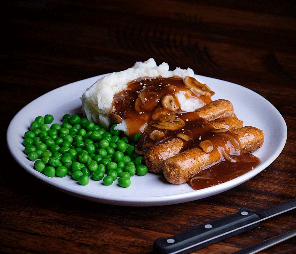 A white plate sits on relative darkness on a dark wooden table. Unreal Co Beefy Brat sausages are covered in gravy, next to a high pile of creamy white mash, with bright pops of green from peas to the side. 