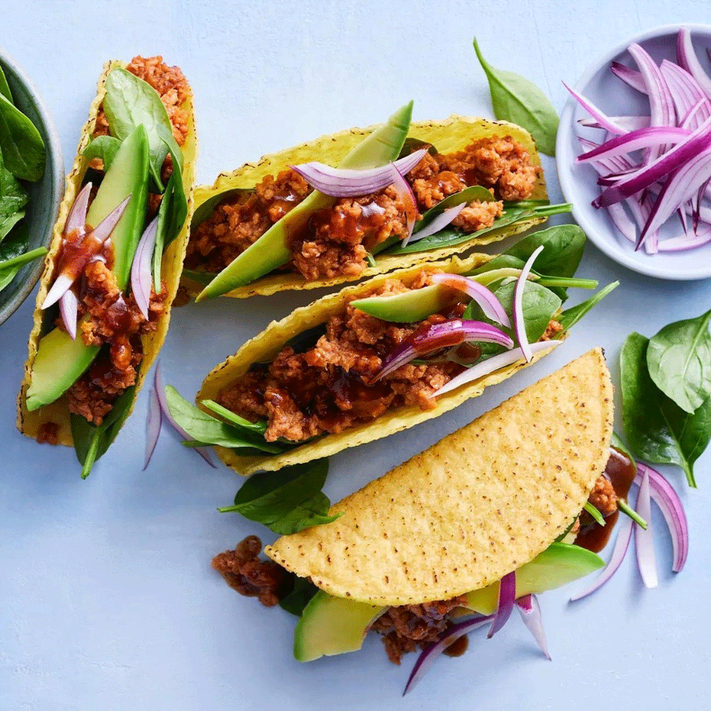 Hard shell tacos sit on a blue surface, filled with V2 plant-based mince, red onion, and avocado slices.