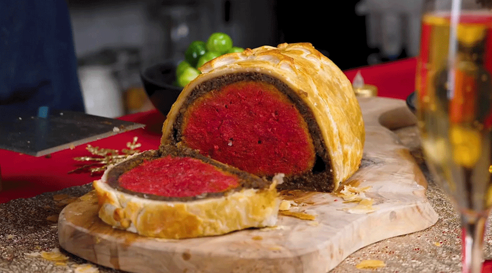 A beef-style wellington sits on a chopping board. It is covered in pastry and a slice has been cut to reveal a red centre, surrounded by a darker filling.