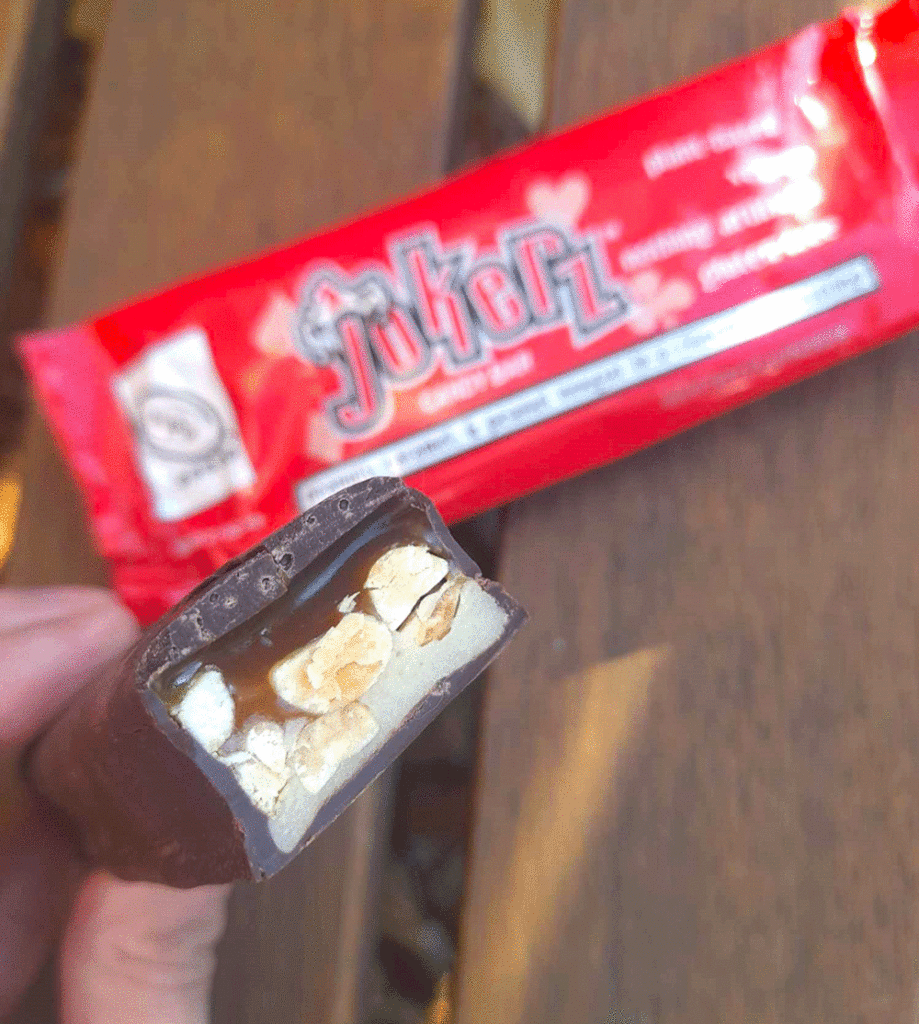 A hand holds a Jokerz chocolate bar which is cut to reveal a cross section of nougat, peanuts and caramel.