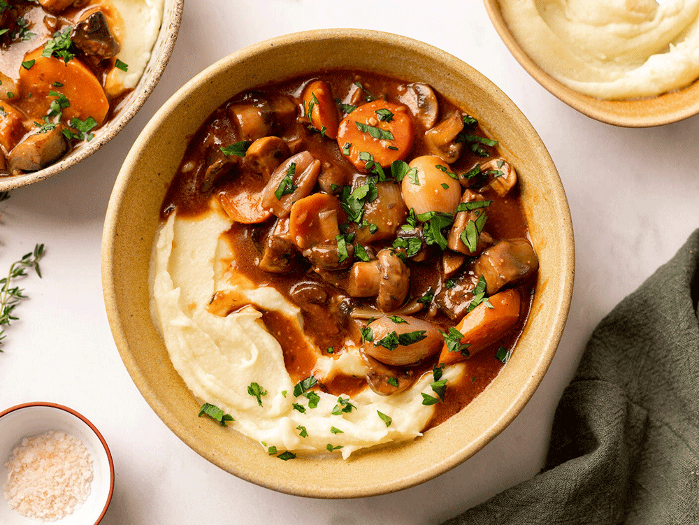 An overhead image of a hearty bowl of mash and beef bourguignon, laden with mushroom, carrot, gravy and shallots. 