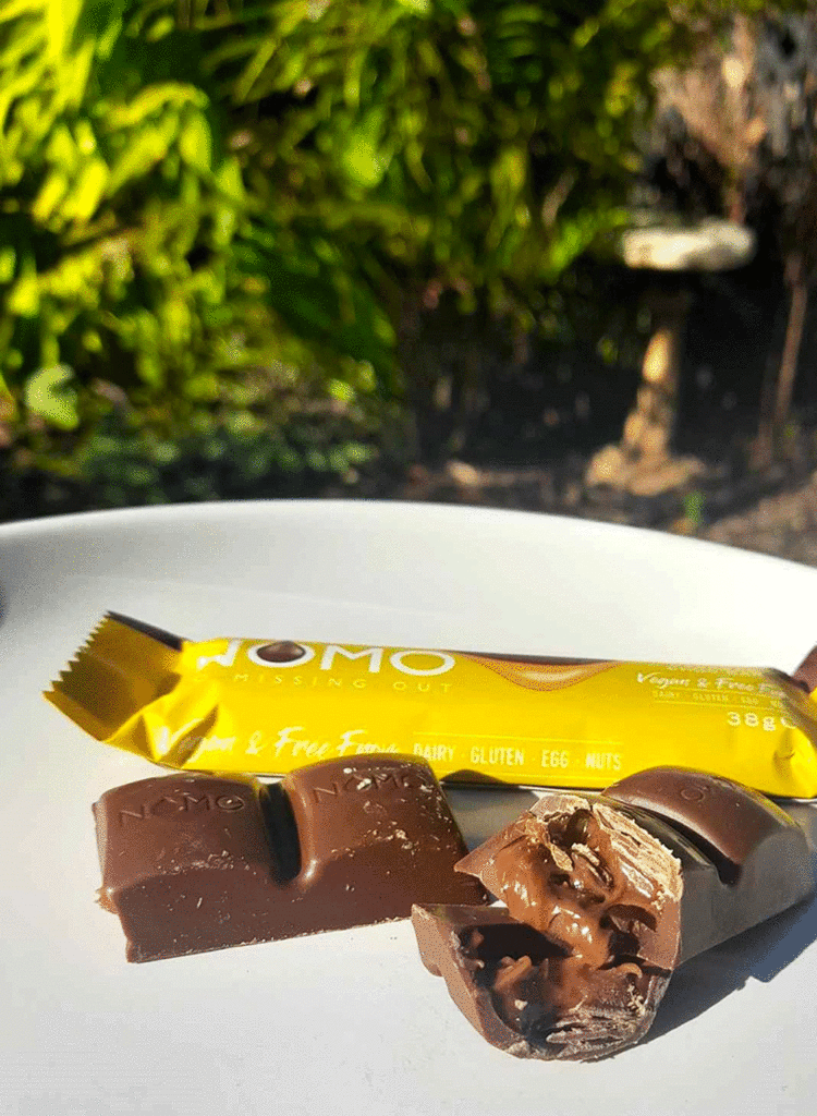 A NOMO bar filled with caramel is broken on a plate. The gooey caramel is glistening in the sun.