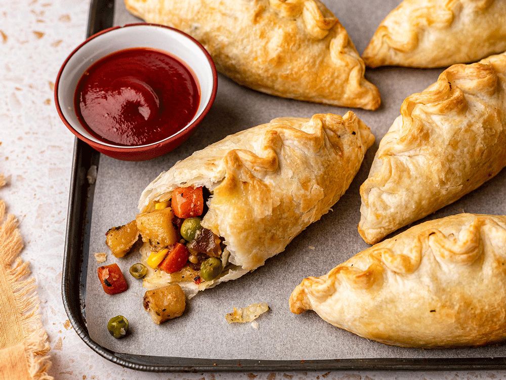 Vegetable pasties sit on a tray, one cut open to reveal a filling of hearty – mainly root – veg. A ramekin of sauce sits to the side.