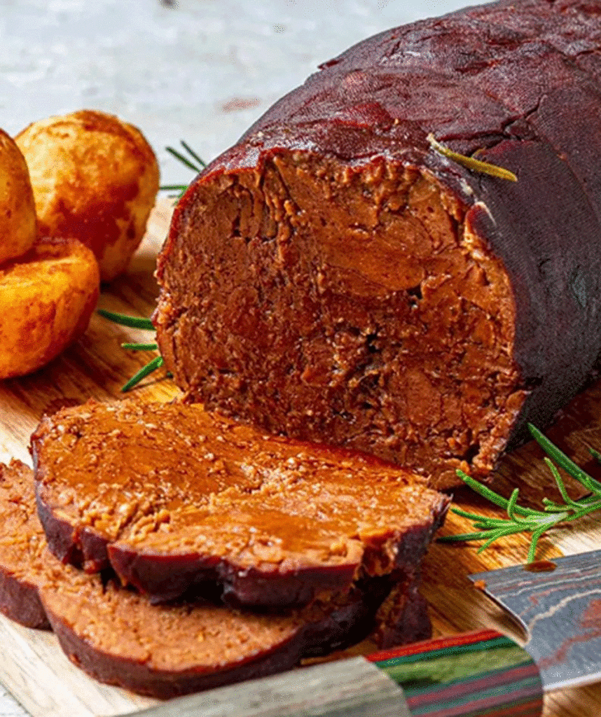 A roast loaf sits on a chopping board, with roast potatoes to the side and decorated with rosemary. A slice is cut revealing a dense, very beefy texture, achieved using seitan. 