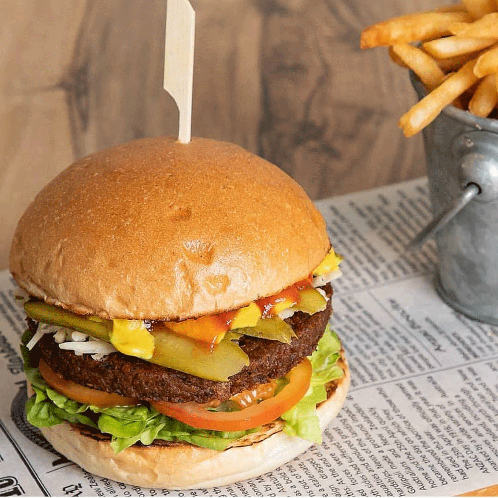 A vEEF plant-based beef burger with all the trimmings sits on a piece of newspaper. A metal bucket of hot chips sits to the side.