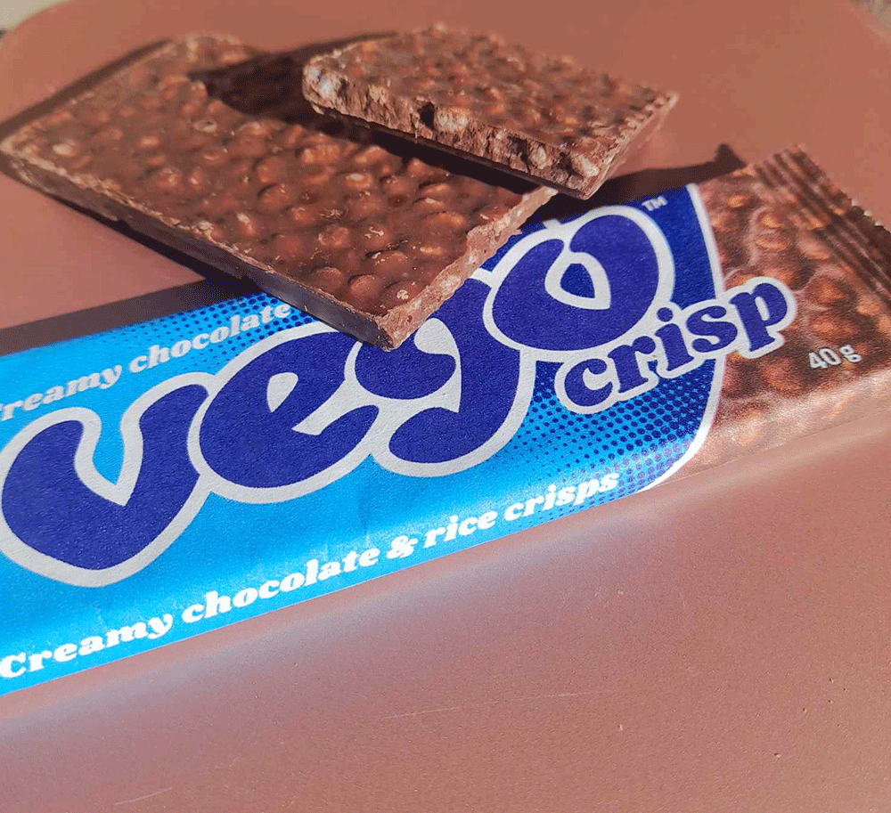 A Vego Crisp sits on a plate, with an open chocolate bar leaning on is, showing the texture of the chocolate rice crisp bar.