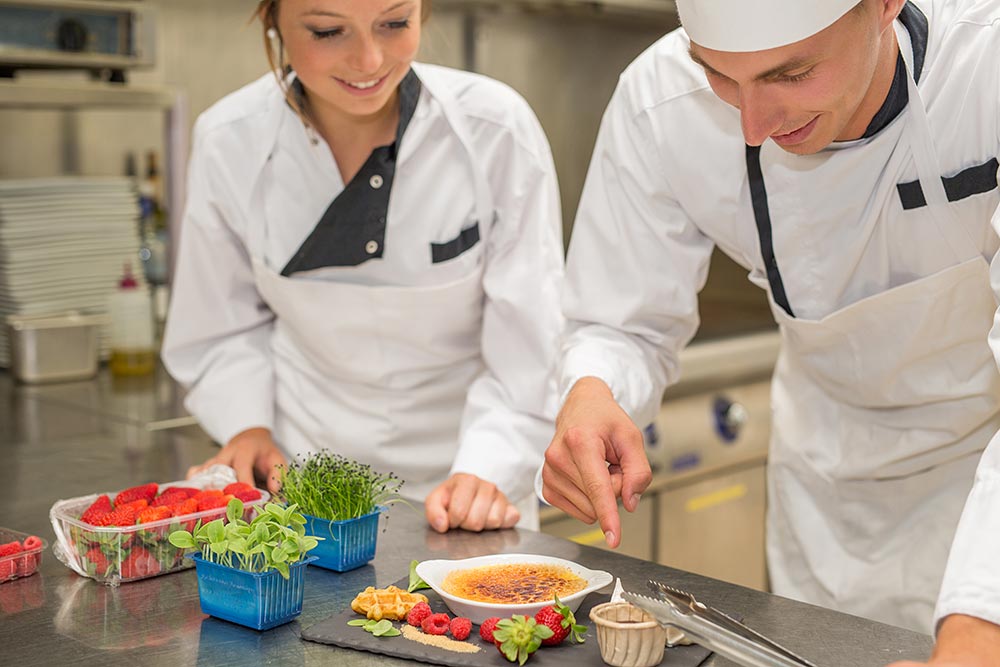 Two chefs work on a bench delicately preparing a dish featuring seasonal fruit and fresh herbs.