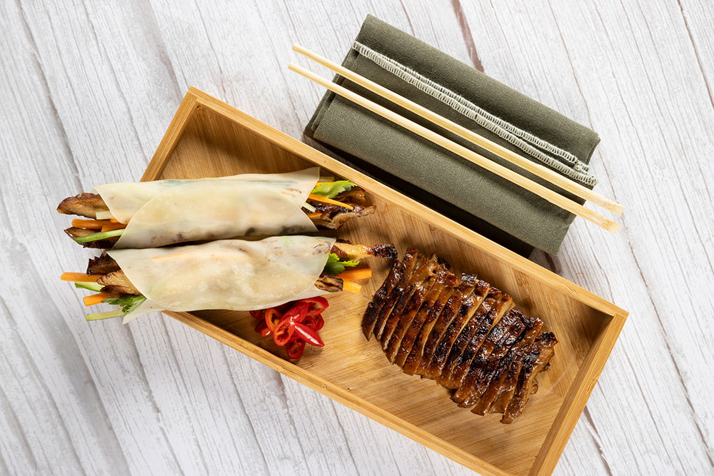 An overhead image of a nicely presented Asian-style roast 'duck', served as both a main and in pancakes on the one wooden serving tray.