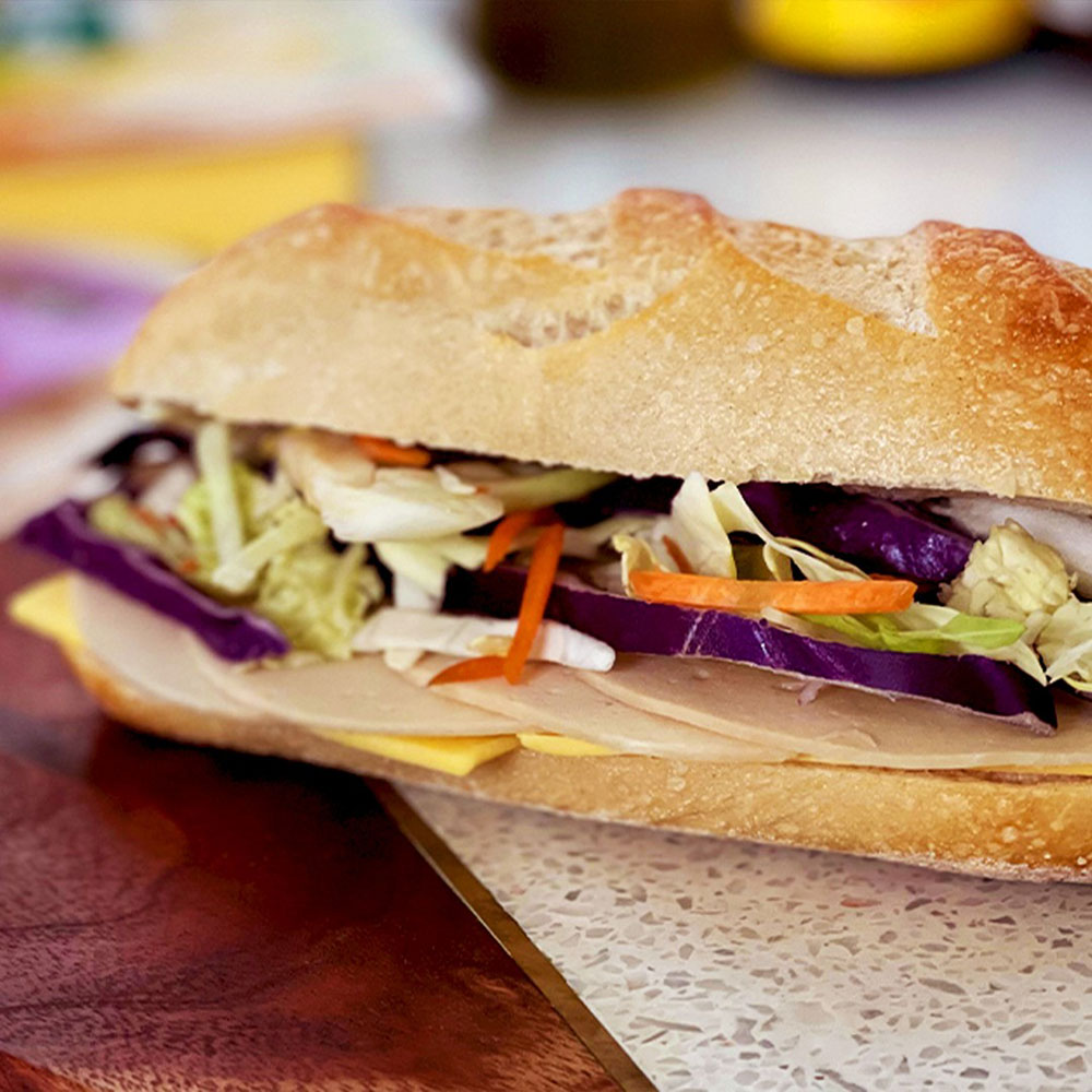 A close up of a baguette filled with Meliora turkey-style deli slices, cheese and slaw.