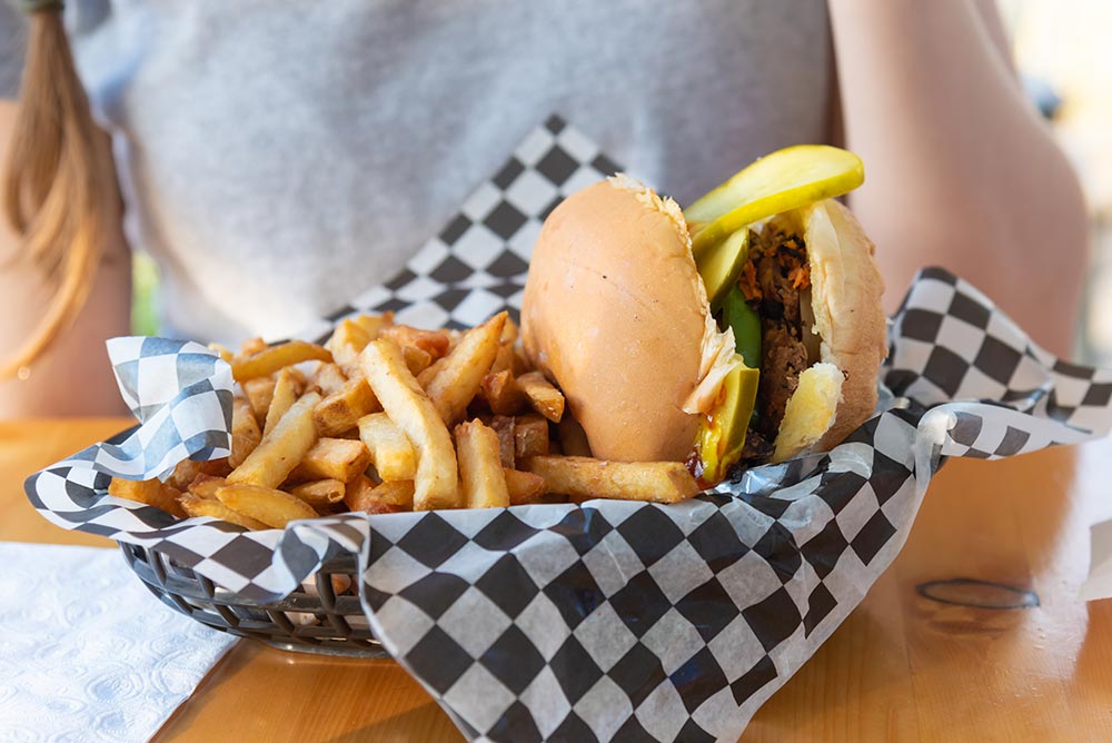 A basket of fries with a plant-based burger sits on a table in front of a woman wearing a grey tshirt.