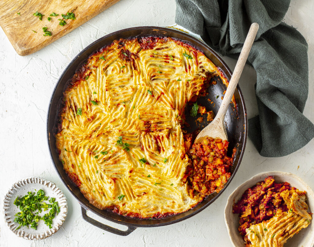 An overhead shot of Lentil Shepherd's pie in an oven dish, with a serve taken out beside it in a bowl. A wooden spoon sits next to the remainder of the pie.