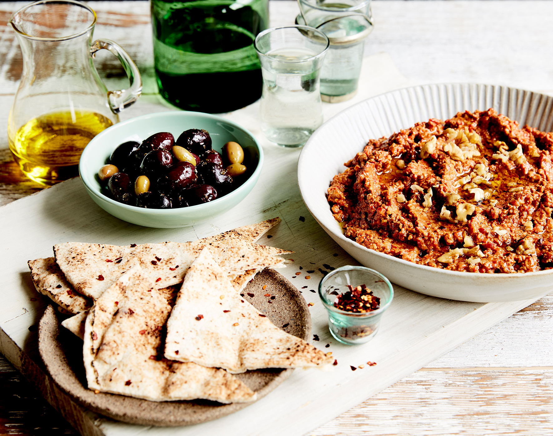 A chunky red dip sits to the left of a table setting, surrounded by pita bread and olives.