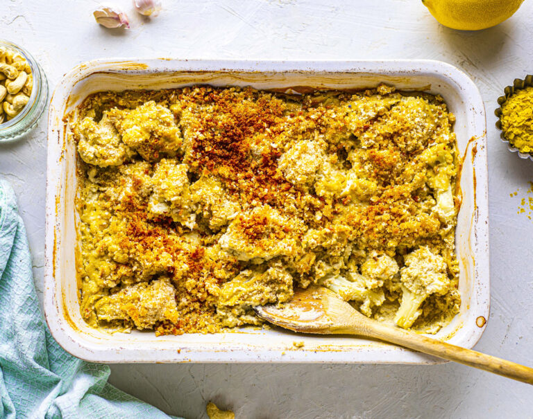 A rectangular oven tray filled with a very creamy looking cauliflower bake. It's topped with breadcrumbs. A wooden spoon sits in the corner.