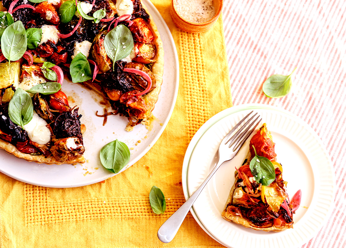 A round tart on a plate with a slice taken out, sitting on a smaller plate next to it. The top is decorated colourfully with tomato and leaves of basil.