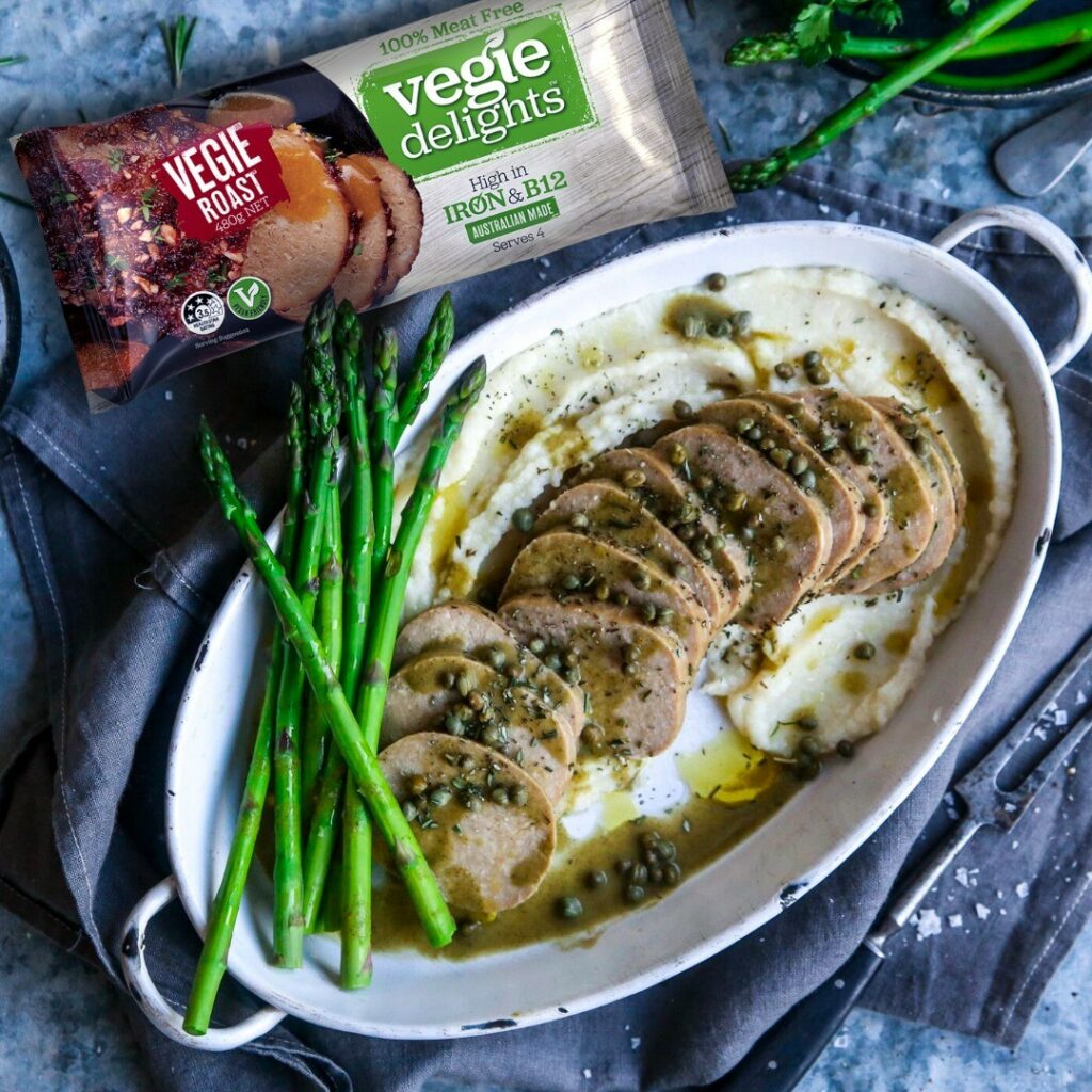 A plant-based roast served up in a baking dish, decorated with asparagus. A Vegie Delights packet sits just in shot..