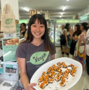 Friendly staff member from the Vegan Grocery Store holds up a tray of food samples instore, smiling.