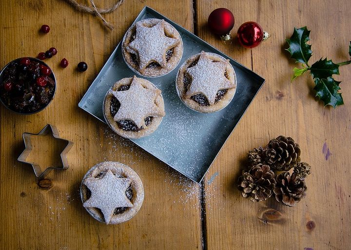 A small tray of 4 mince pies on a table surrounded by Christmas decorations.