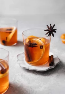 Three glasses of cider sit on a table. The central glass is on a saucer and all are filled with mulled cider. They are garnished with orange slices and star anise.