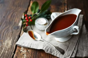 A gravy boat full of rich gravy on a timber table, atop a fabric napkin and some xmas holly.