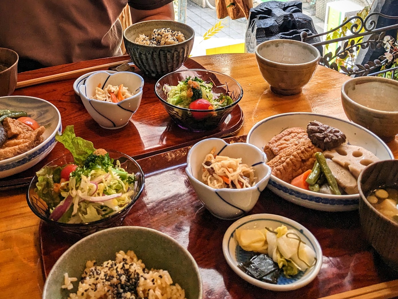 A cosy, wooden cafe table peppered with small bowls of various shapes and styles. Each bowl holds a different macrobiotic-style Japanese food such as pickled veggies, seaweed, lotus root, bean curd, mushrooms, brown rice, miso soup, and tofu.