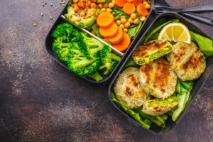 Healthy meal prep containers with green burgers, broccoli, chickpeas and salad on dark background.