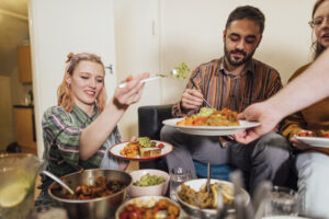 Group of student house mates sitting around a table eating together.They have cooked creole aubergines and quinoa with sweet potatoes, peppers and avocado puree.