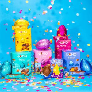 A range of NOMO Easter products sit colourfully in front a sky blue background, with confetti falling around them.