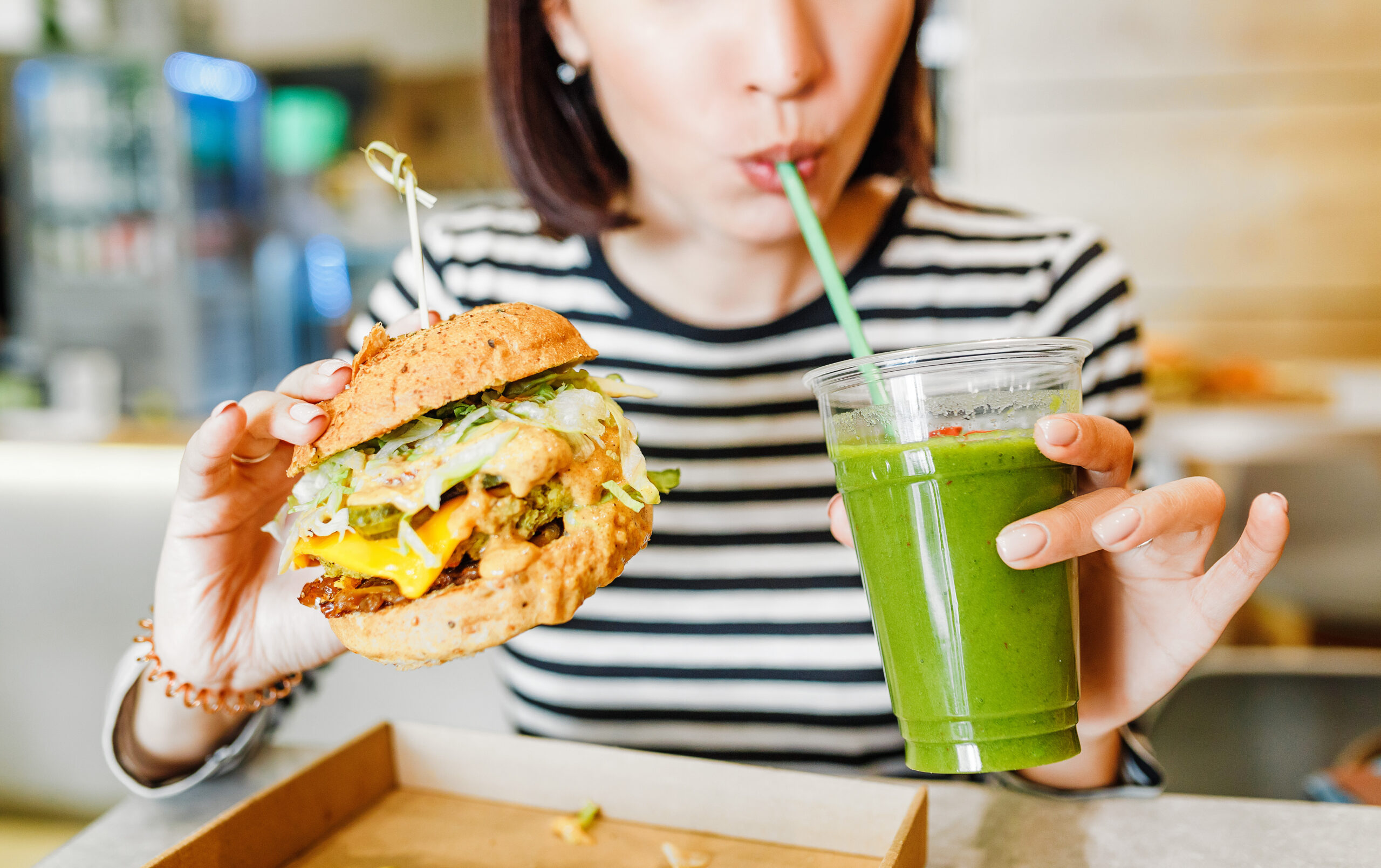 A young woman drinks a green smoothie and eats a burger in a vegan fast food restaurant