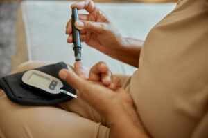 Diabetes patient checks their sugar level with a needle prick in their finger, while a glucometer sits on their knee.