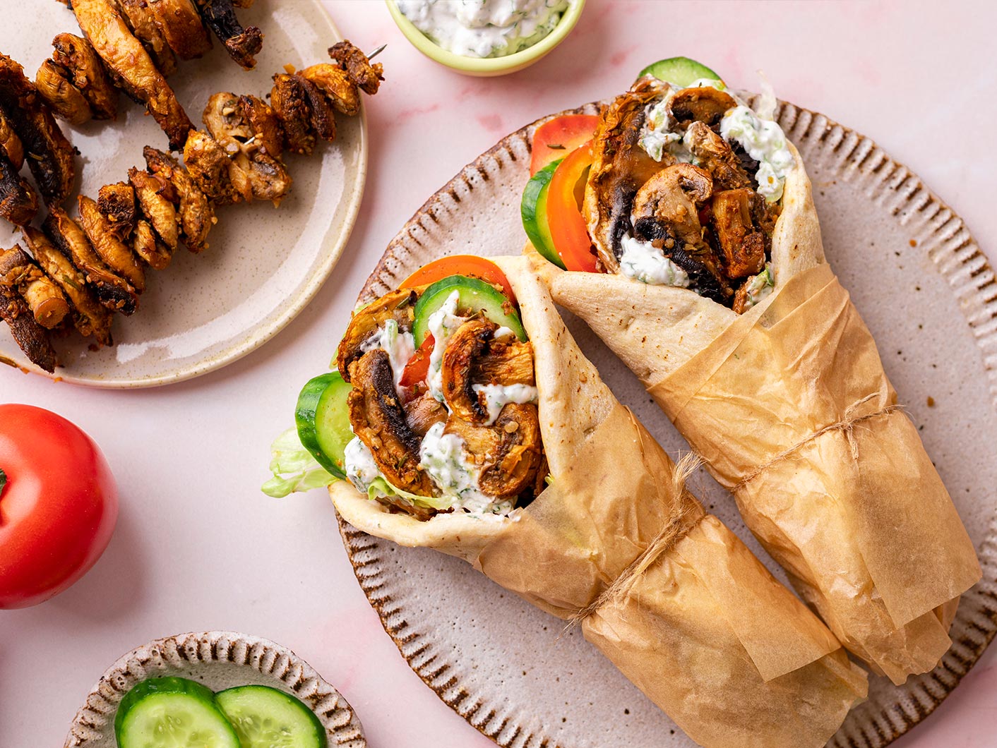 Mushroom souvlaki served up with salad and tzatziki on a light coloured plate. Mushroom skewers sit on a plate just in shot, on a light pink bench.