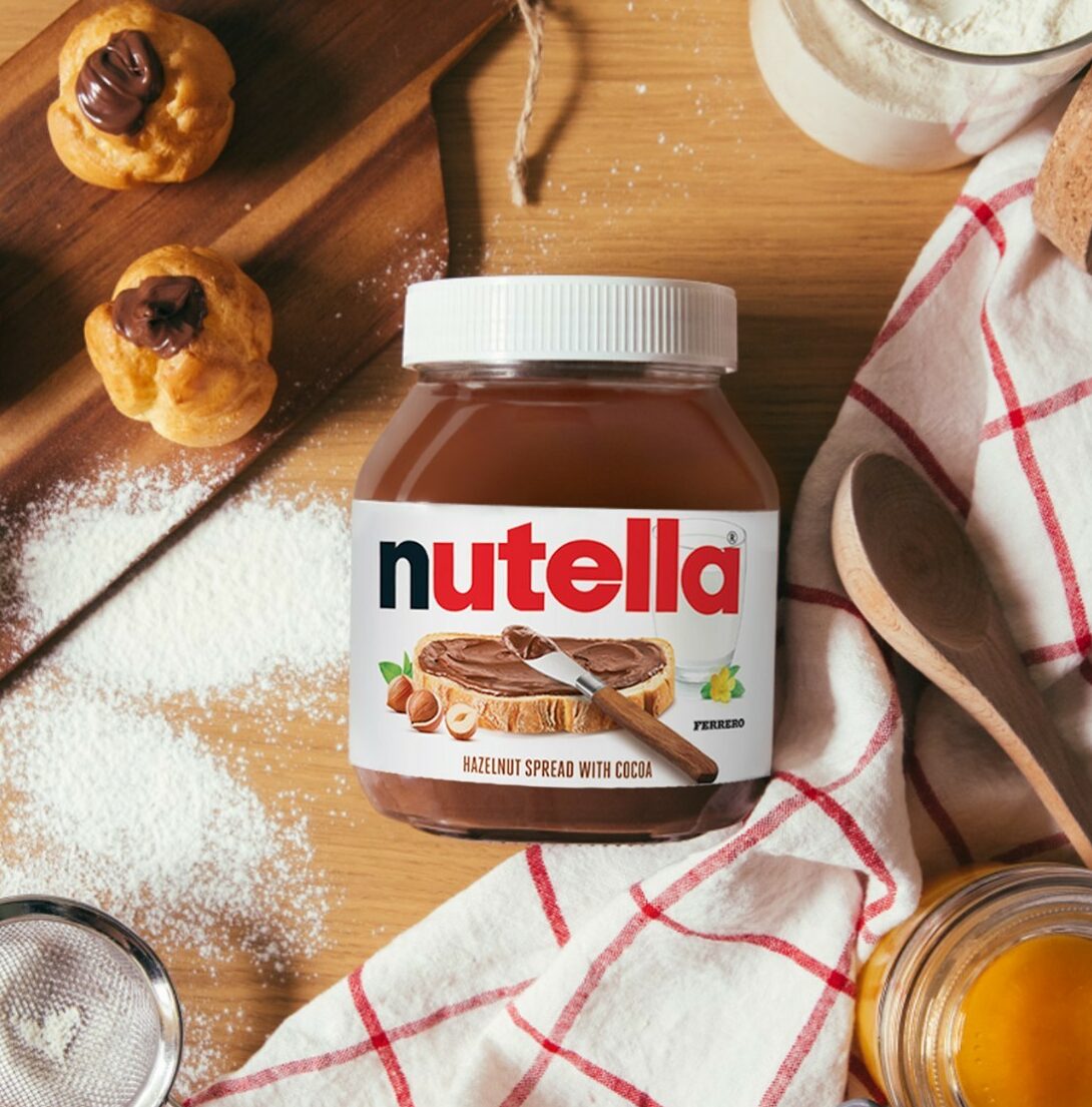 Overhead image of a jar of Nutella on a wooden bench,. Around it sits some Nutella-filled profiteroles, a tea towel, wooden spoon, and icing sugar is dusted on the bench.