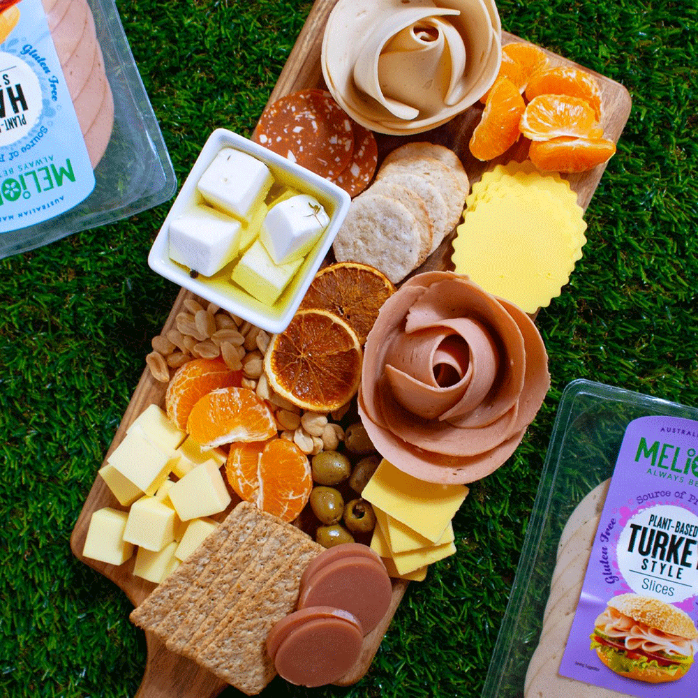 A colouful and beautifully presented charcuterie board sits on green grass. It features crackers, an assortment of fruits and plant-based cheeses, and plant-based deli slices in ham and turkey-style, prepared into roses.